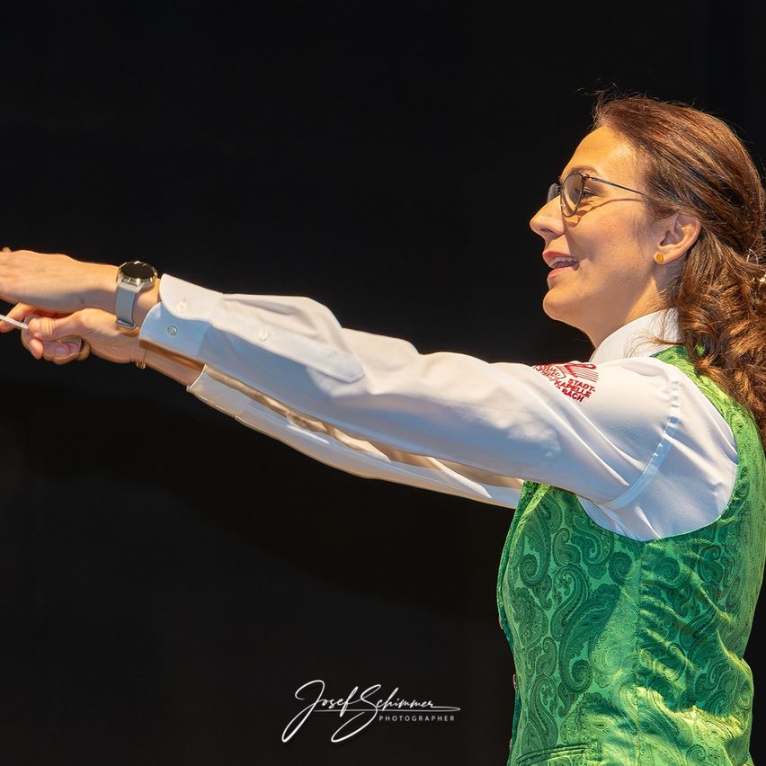 Eine Frau dirigiert ein Orchester, trägt eine grüne Weste und hält einen Taktstock. Sie lächelt mit geschlossenen Augen.