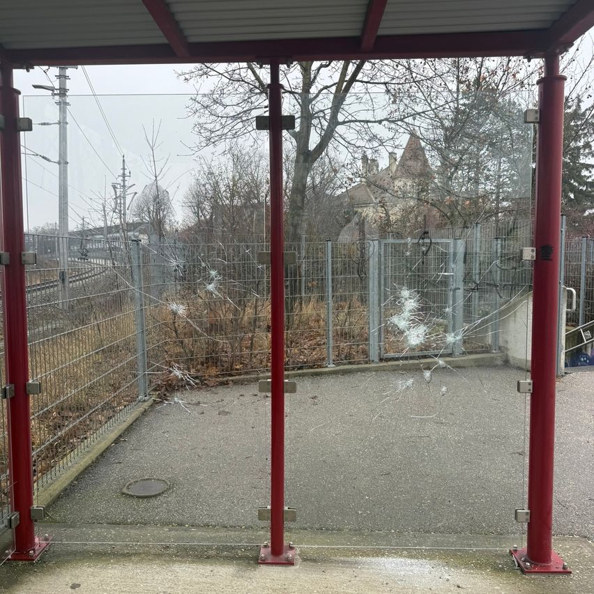 A bus stop with broken glass, red poles, and a metal fence. The area is surrounded by trees and a building.