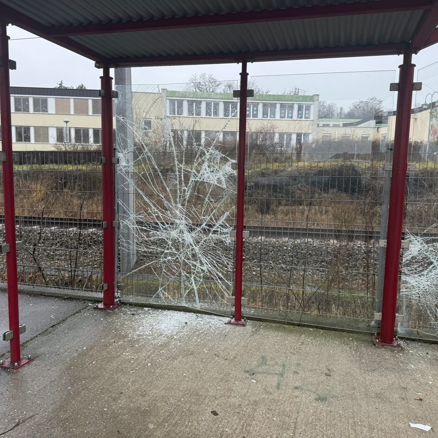 A bus stop with a cracked glass window and metal fence, near a school building and a train track.