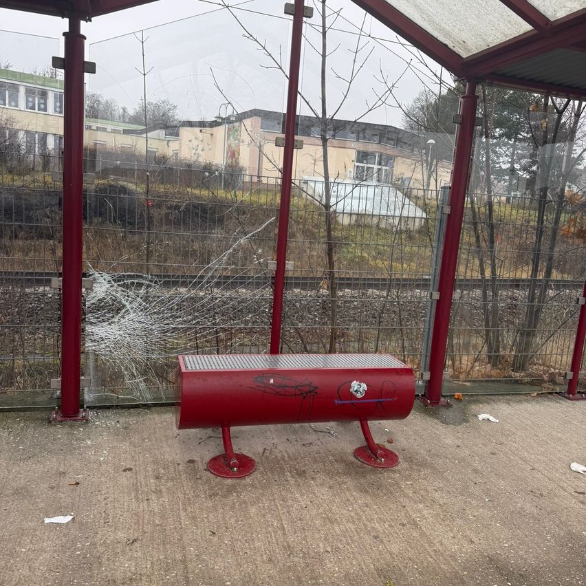 A damaged red bench is at a bus stop with a cracked glass panel. It is outdoors and surrounded by a metal fence. There are trees and a building in the distance.