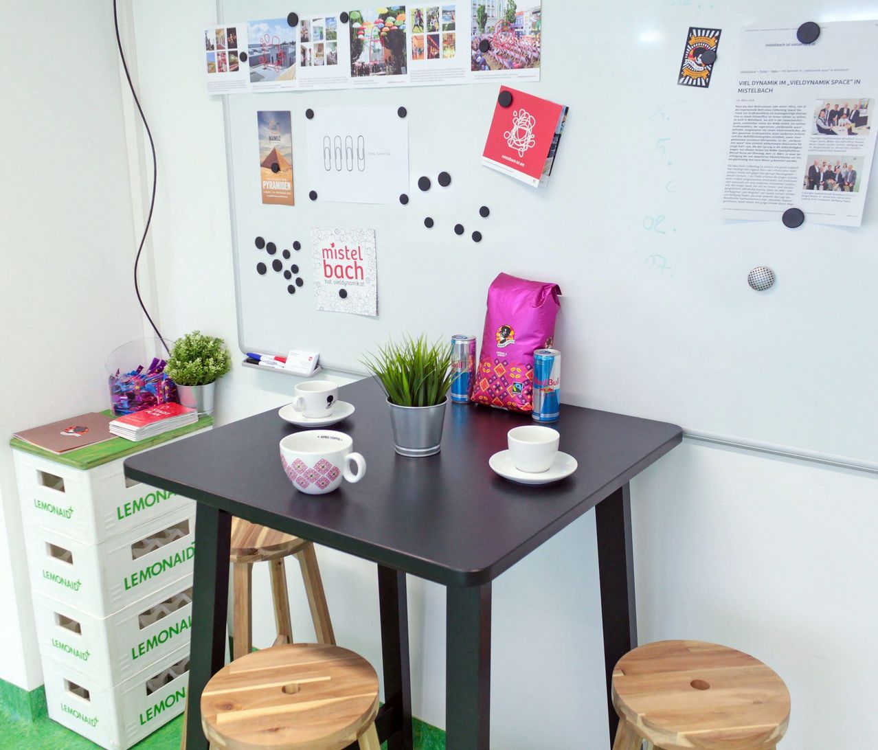A small table with two cups and saucers, a potted plant, and a bag sits in front of a white wall with various items attached.