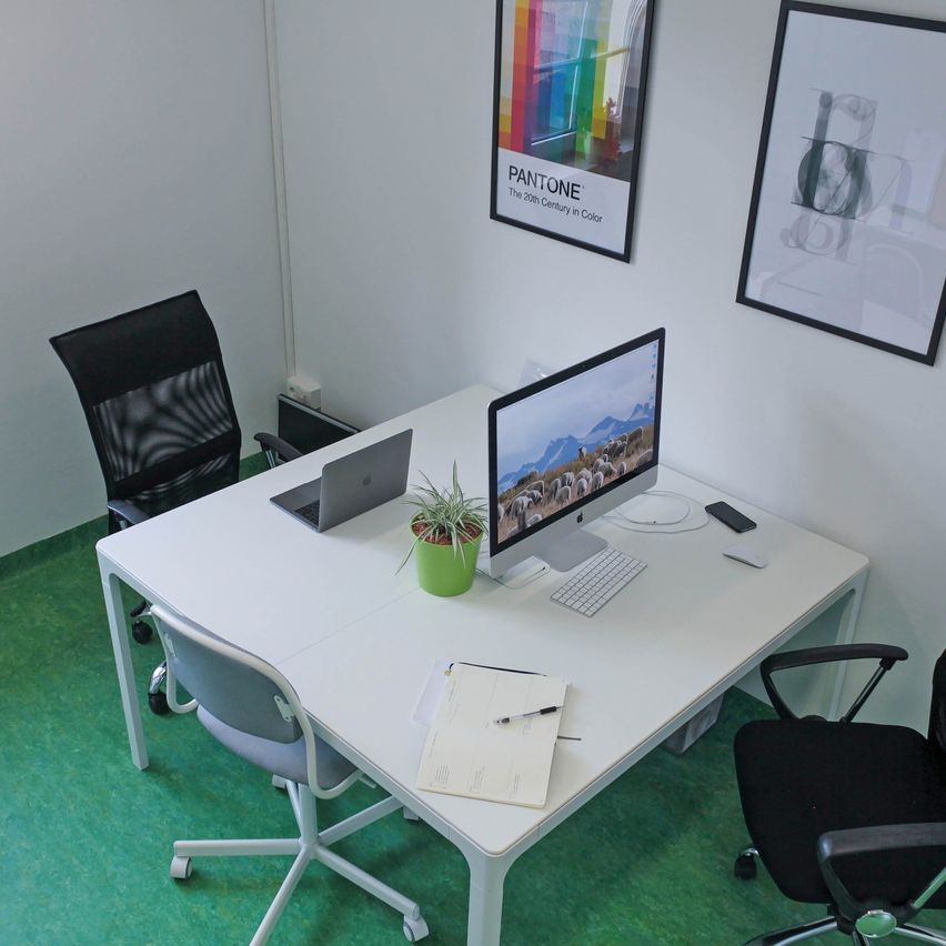 An office with a white desk, two chairs, a potted plant, a monitor, keyboard, mouse, and papers on the desk. A poster on the wall reads 'The 20th Century in Color'.