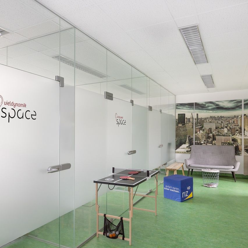 A modern office with glass walls features a ping-pong table and a gray couch. The room includes a blue storage box and a city mural.