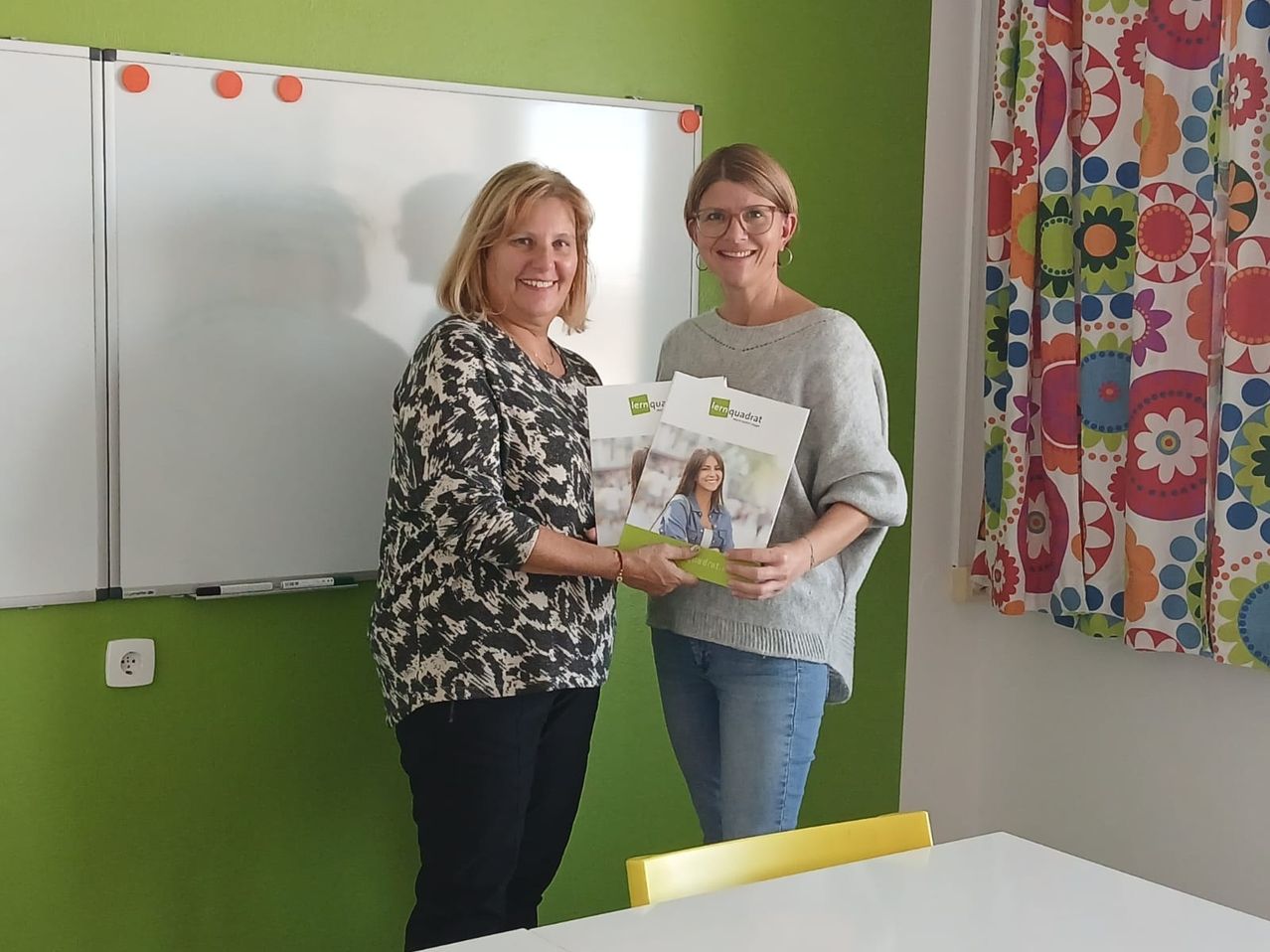 Zwei Frauen stehen nebeneinander und halten Broschüren vor einer grünen Wand und einer Tafel. Eine trägt ein Blumenhemd, die andere ein graues Sweatshirt.