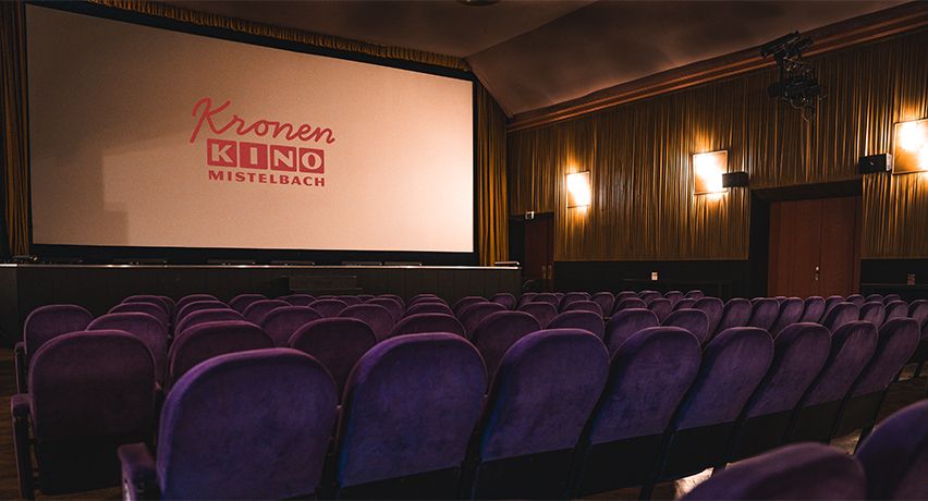 The interior of a cinema theater named Krone Kino Mistelbach, showing rows of purple chairs, a large screen, and ambient lighting.