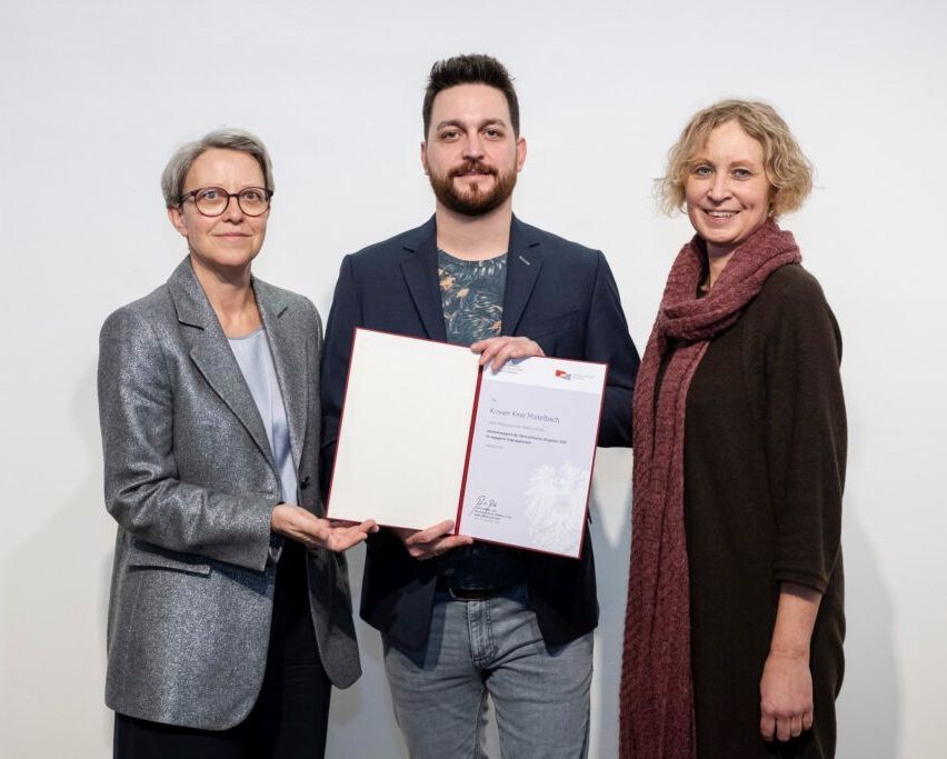 Three people pose for a photo with a certificate, the man in a suit holds it in both hands. The woman on the left wears glasses and a gray blazer. The woman on the right smiles and wears a scarf.