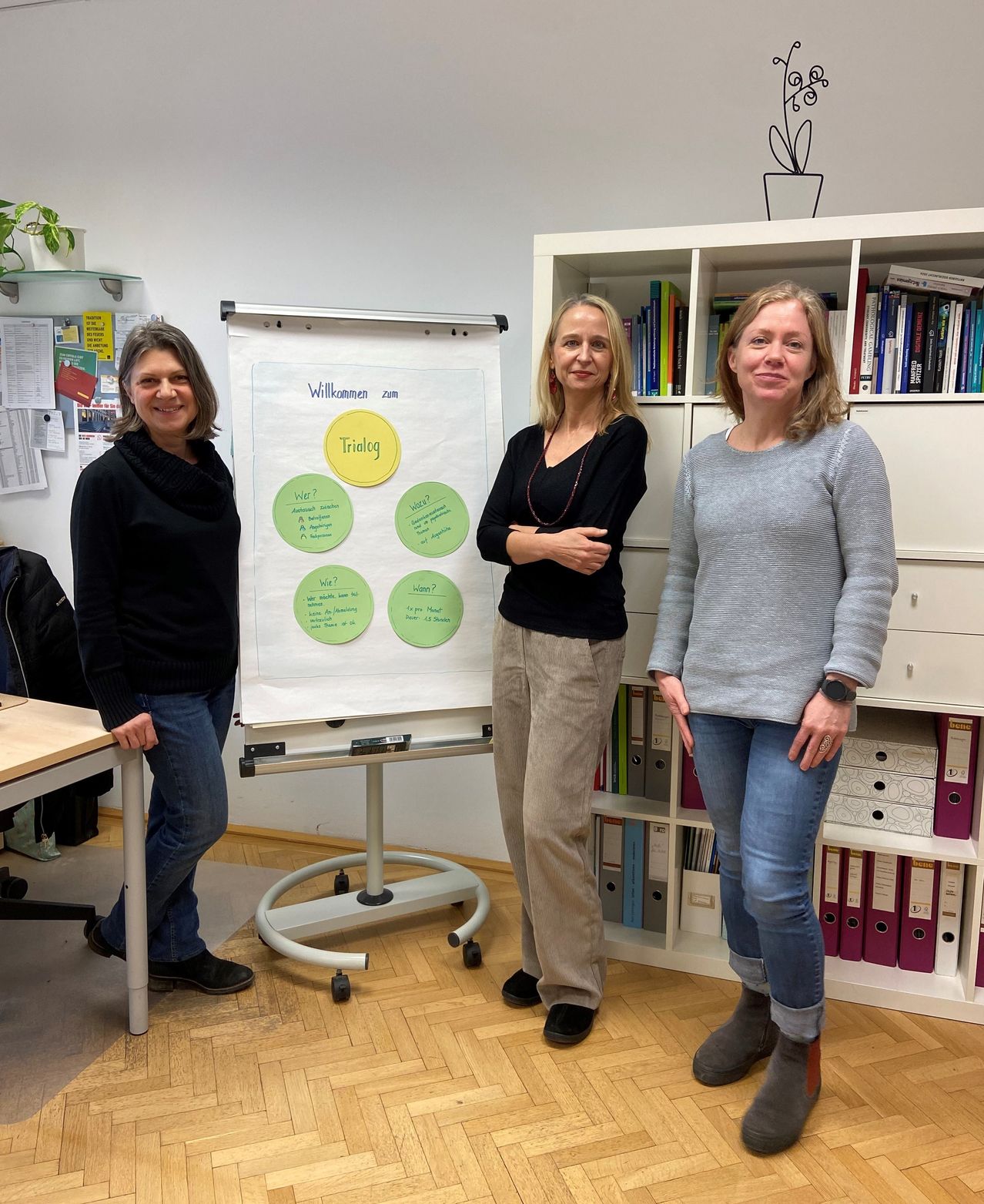 Three women pose for a photo in an office, standing by a whiteboard and a bookshelf. The woman on the left wears a black sweater and jeans. The center woman wears a black top and khaki pants. The woman on the right wears a gray sweater and jeans.