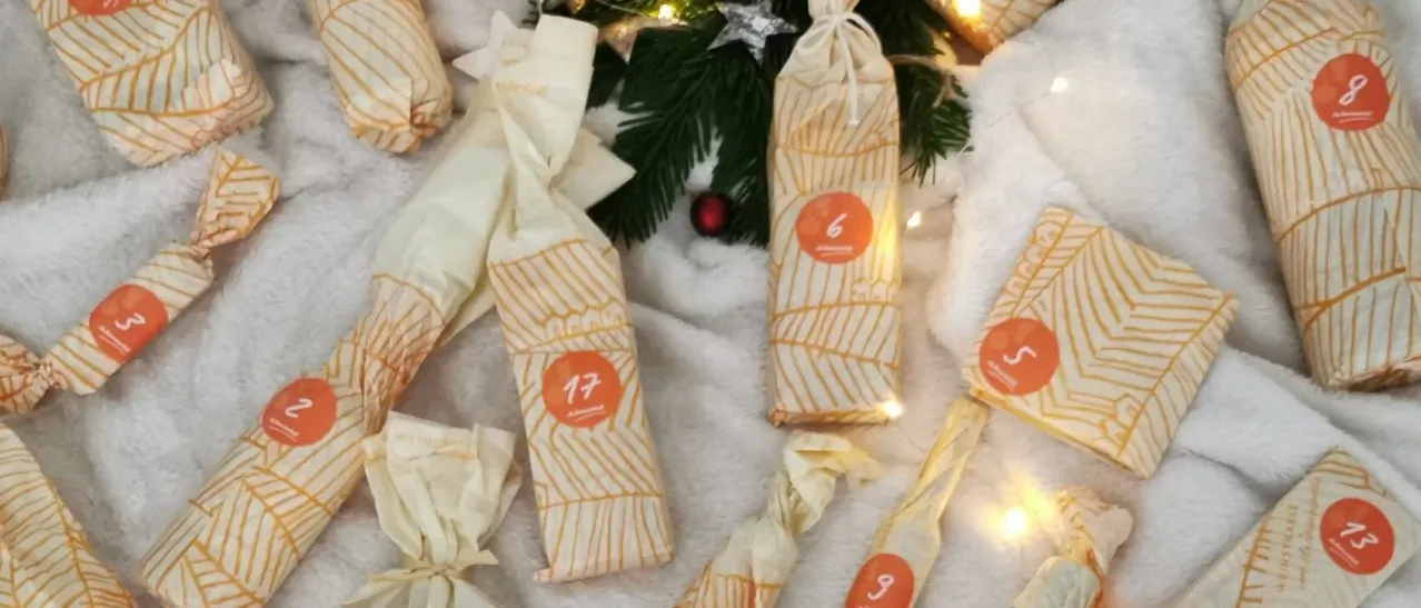Mehrere verpackte Geschenke mit orangefarbenen Nummern sind auf einem weißen Tuch angeordnet, möglicherweise für einen Feiertag, mit einem Weihnachtsbaum im Hintergrund.