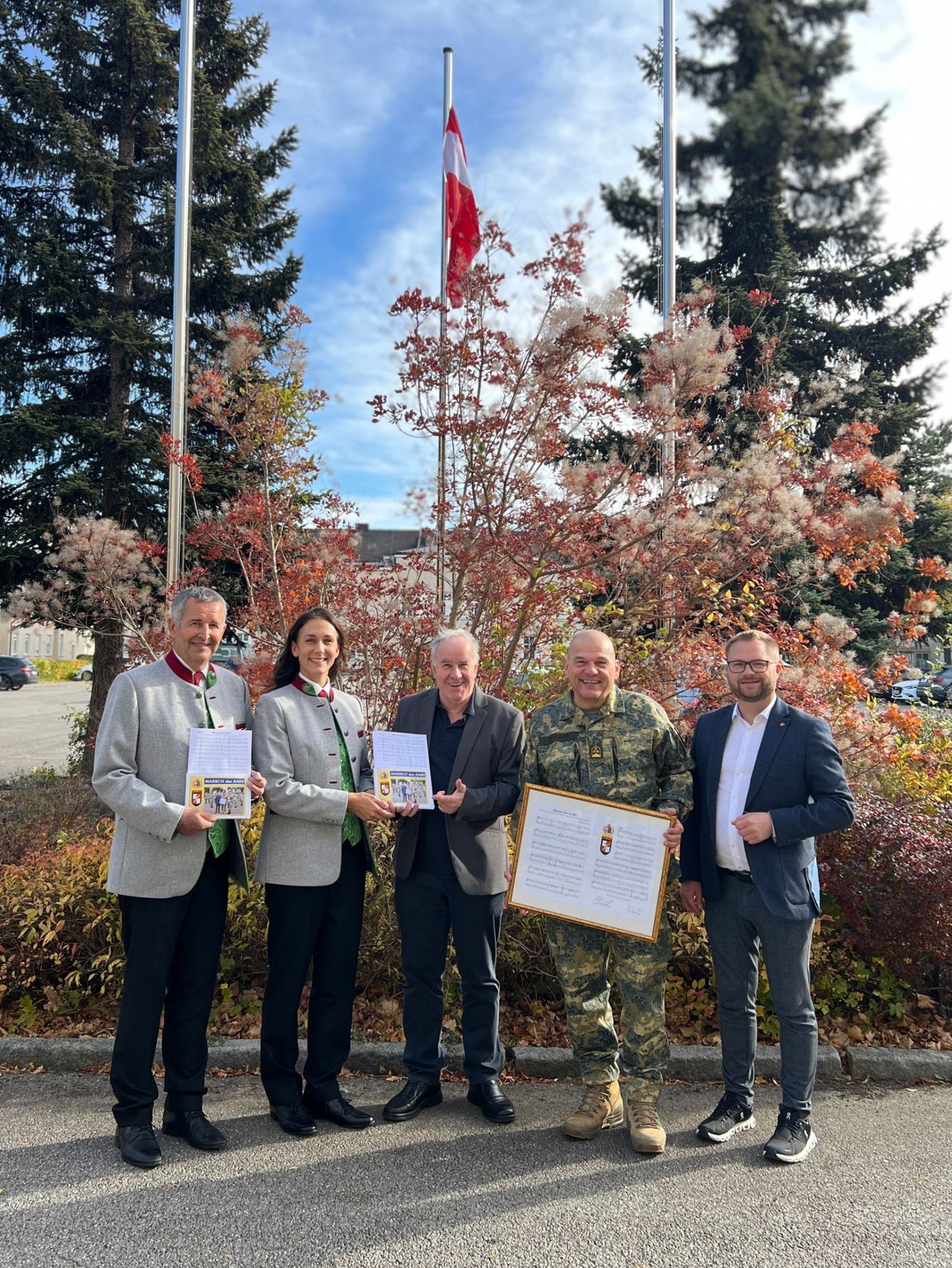 Fünf Personen stehen vor Bäumen und halten Urkunden und ein gerahmtes Dokument in der Hand. Zwei sind in Militäruniformen und drei in Geschäftskleidung. Dahinter sind zwei Fahnenmasten mit Fahnen zu sehen, und ein Gebäude ist in der Ferne.