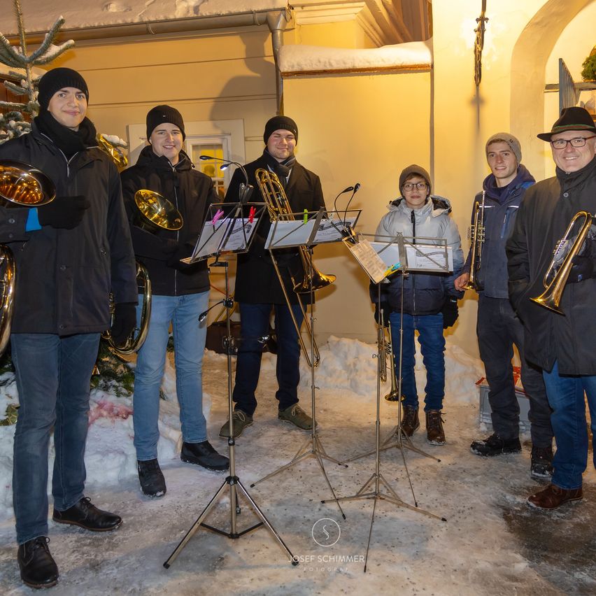 Eine Gruppe von Musikern steht im Schnee mit ihren Instrumenten. Sie sind warm gekleidet, mit Winterkleidung und Mützen. Sie lächeln und scheinen für ein Foto zu posieren.