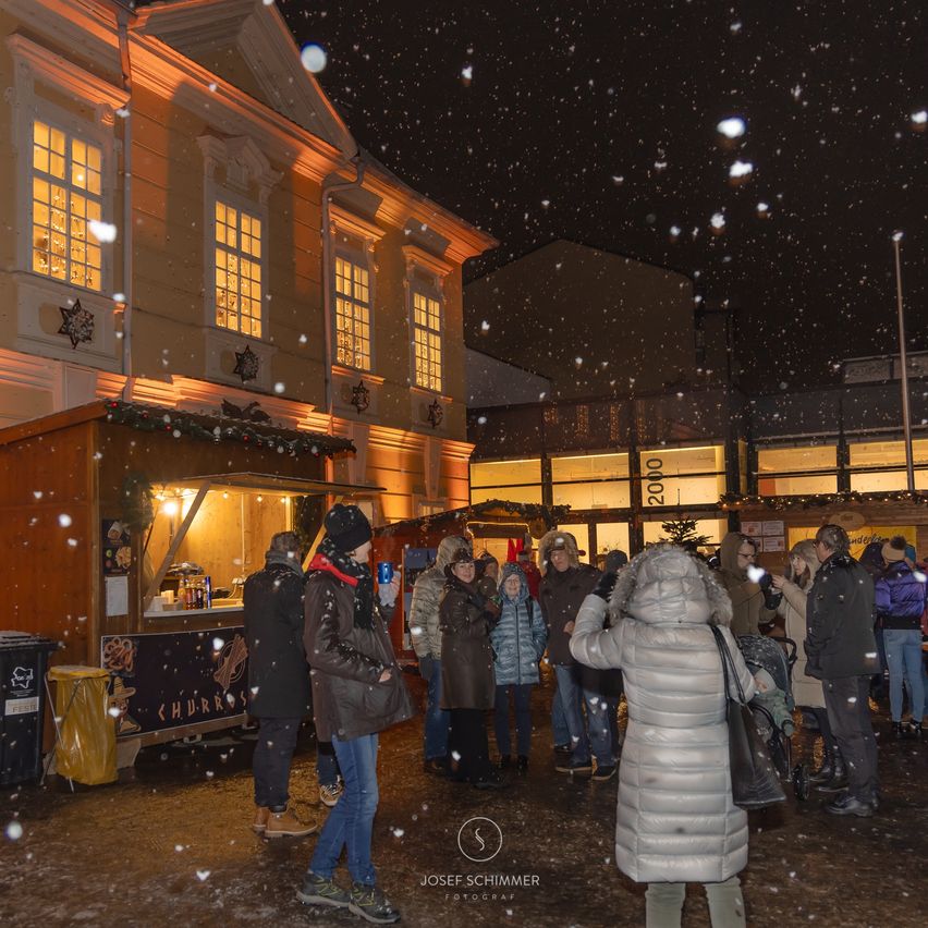 Menschen versammeln sich um einen Imbissstand in einem verschneiten Bereich, tragen Winterkleidung. Eine Frau in einer mit Fell gefütterten Jacke macht ein Foto. Das Gebäude hinter ihnen hat beleuchtete Fenster.