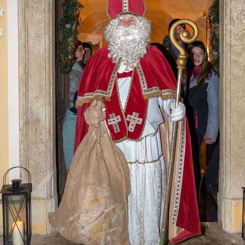 Ein Mann, der als Weihnachtsmann verkleidet ist, steht in einer Tür. Er hält einen Stab und einen Sack. Er trägt einen roten Umhang und einen weißen Gewand. Eine Frau und ein Kind stehen hinter ihm.