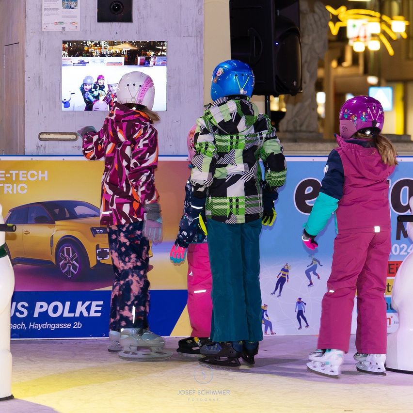 Four children in winter gear are watching a video on a monitor. They stand on an ice rink with a yellow car and a blue banner in the background.