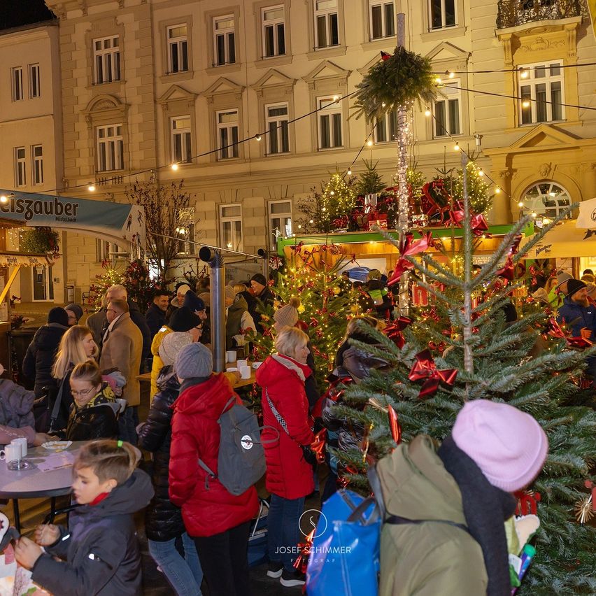 At night, a Christmas market is bustling with people. Decorated trees and stalls are illuminated, and families are browsing and enjoying the festive atmosphere.