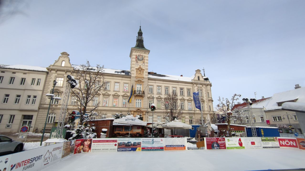 A wintery scene with an ice rink in front of a large historic building, featuring a clock tower, flags, and a blue banner. There are small shops with canopies and various advertisements.