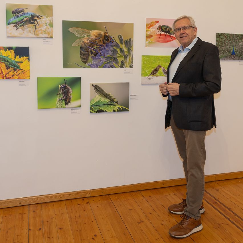 A man stands in front of a white wall with various insect photographs, wearing a black blazer and brown pants.