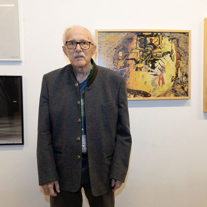 An elderly man stands in front of a white wall, wearing a gray jacket and eyeglasses. Behind him, there are three framed artworks: a yellow tractor, a black-and-white photo, and a white piece.