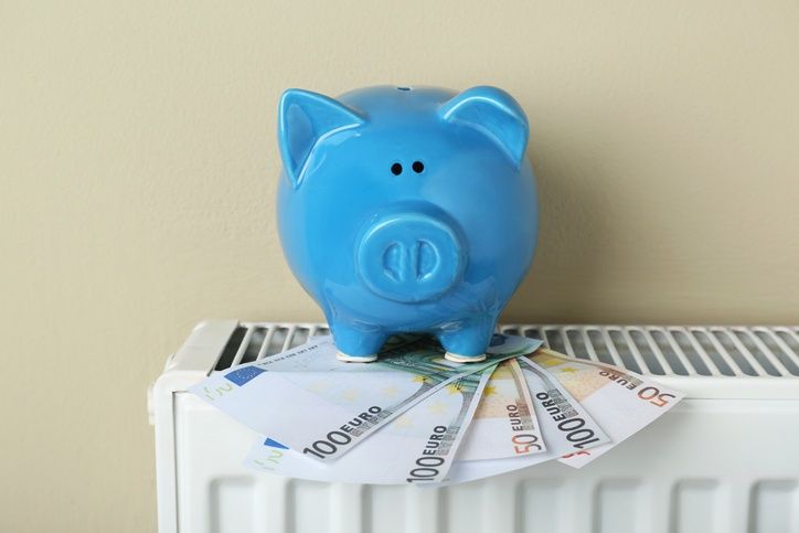 A blue piggy bank with coins sits atop a white radiator against a beige wall. Below it, various Euro bills are stacked.