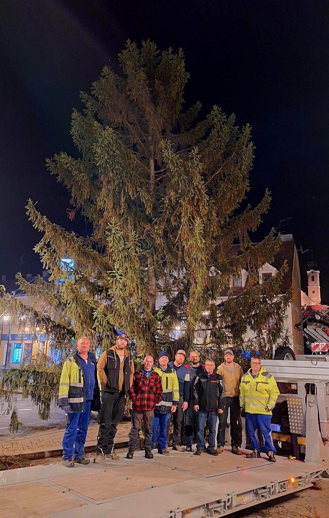 A group of people stand under a large fir tree at night, dressed in winter clothes and some with hats. They appear to be posing for a photograph.