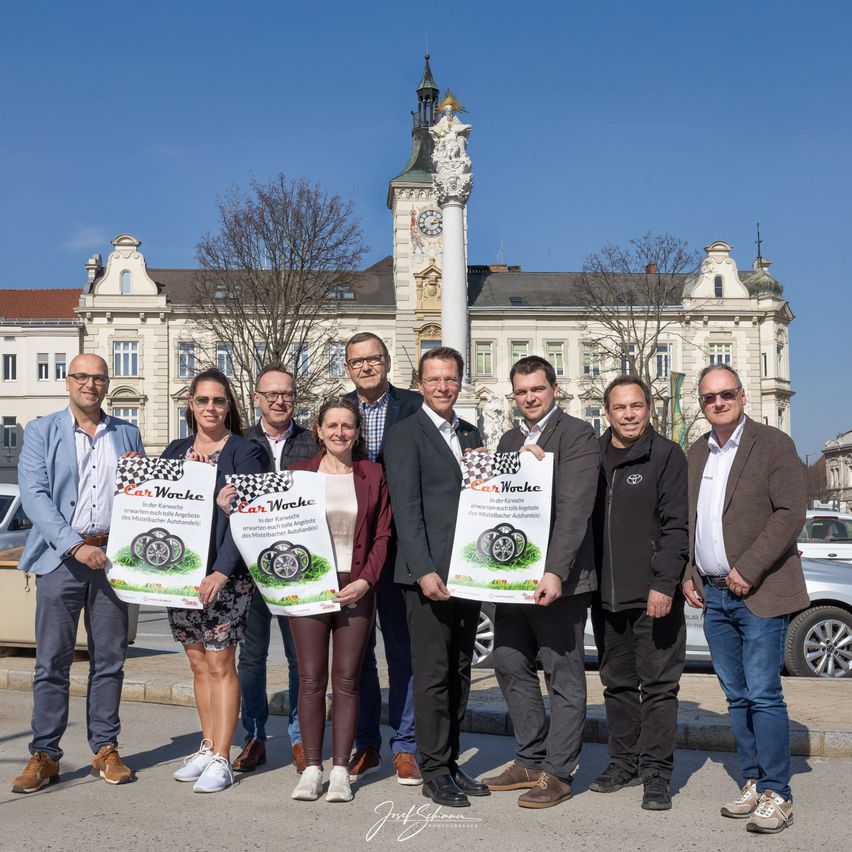 Eine Gruppe von Menschen mit Schildern steht vor einem Gebäude mit Uhrenturm. Sie lächeln und posieren für ein Foto. Das Gebäude ist weiß mit einigen Fenstern.