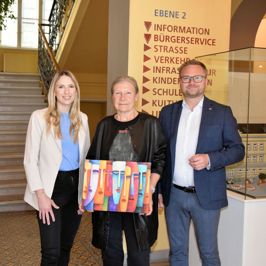 Drei Personen stehen im Innenbereich; zwei Frauen und ein Mann, lächelnd und posieren für ein Foto. Die Frau auf der linken Seite trägt einen weißen Blazer und ein blaues Oberteil, während die Frau in der Mitte ein farbenfrohes Gemälde hält. Der Mann auf der rechten Seite trägt einen dunklen Anzug. Eine Treppe und eine gelbe Wand mit Text stehen hinter ihnen.