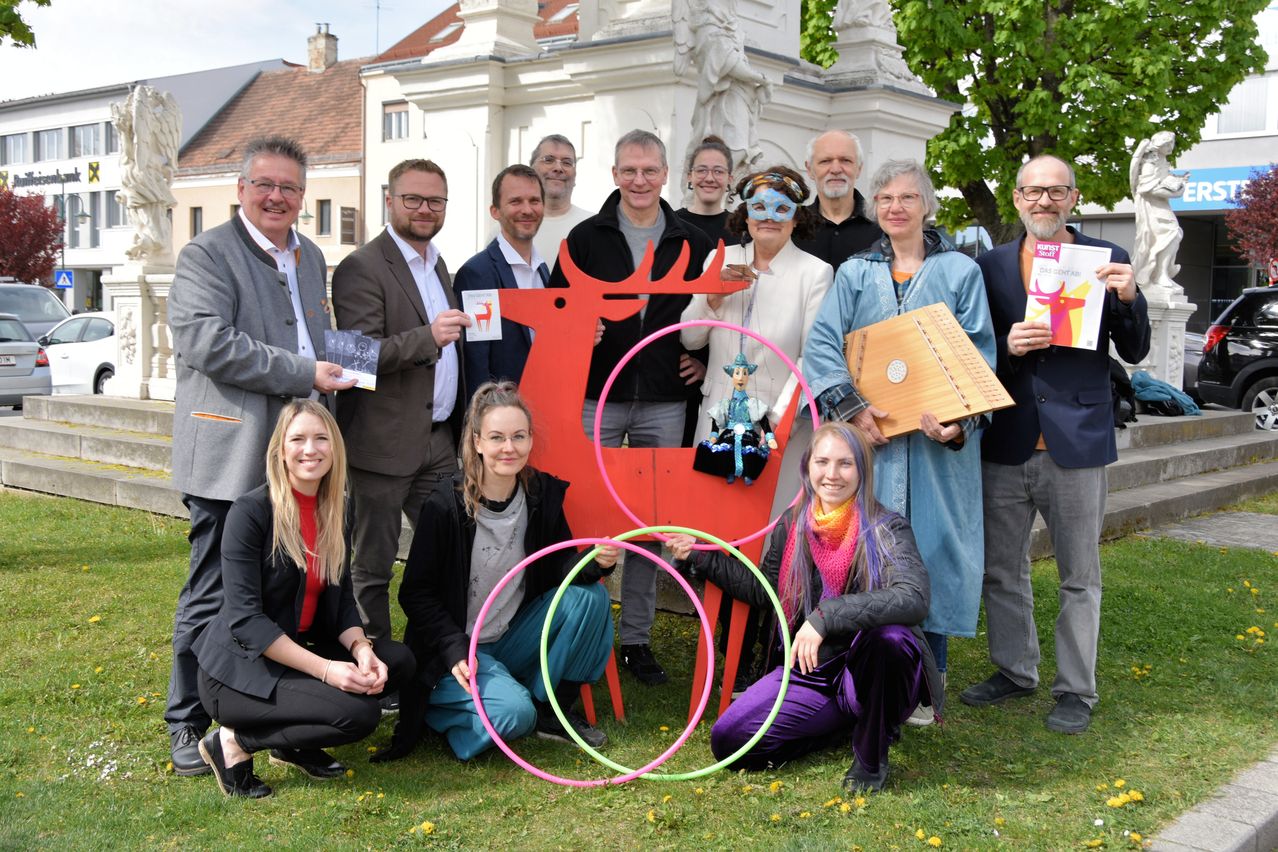 Eine Gruppe von Menschen posiert für ein Foto mit einer großen roten Rentier-Skulptur, hält verschiedene Gegenstände in den Händen und trägt Freizeitkleidung in einem Park.