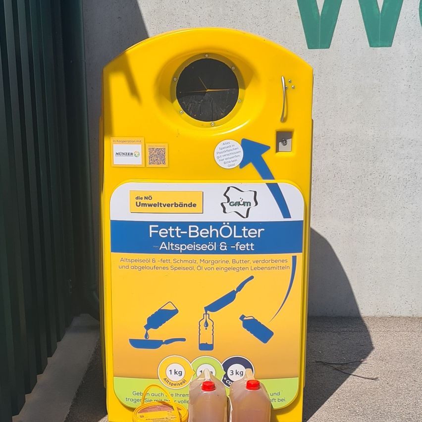 Ein gelber Container mit der Aufschrift Fett-Behölter Altspeiseöl & Fett. Zwei volle Ölflaschen stehen neben ihm. Das Etikett enthält Anweisungen und Gewichtsgrenzen.
