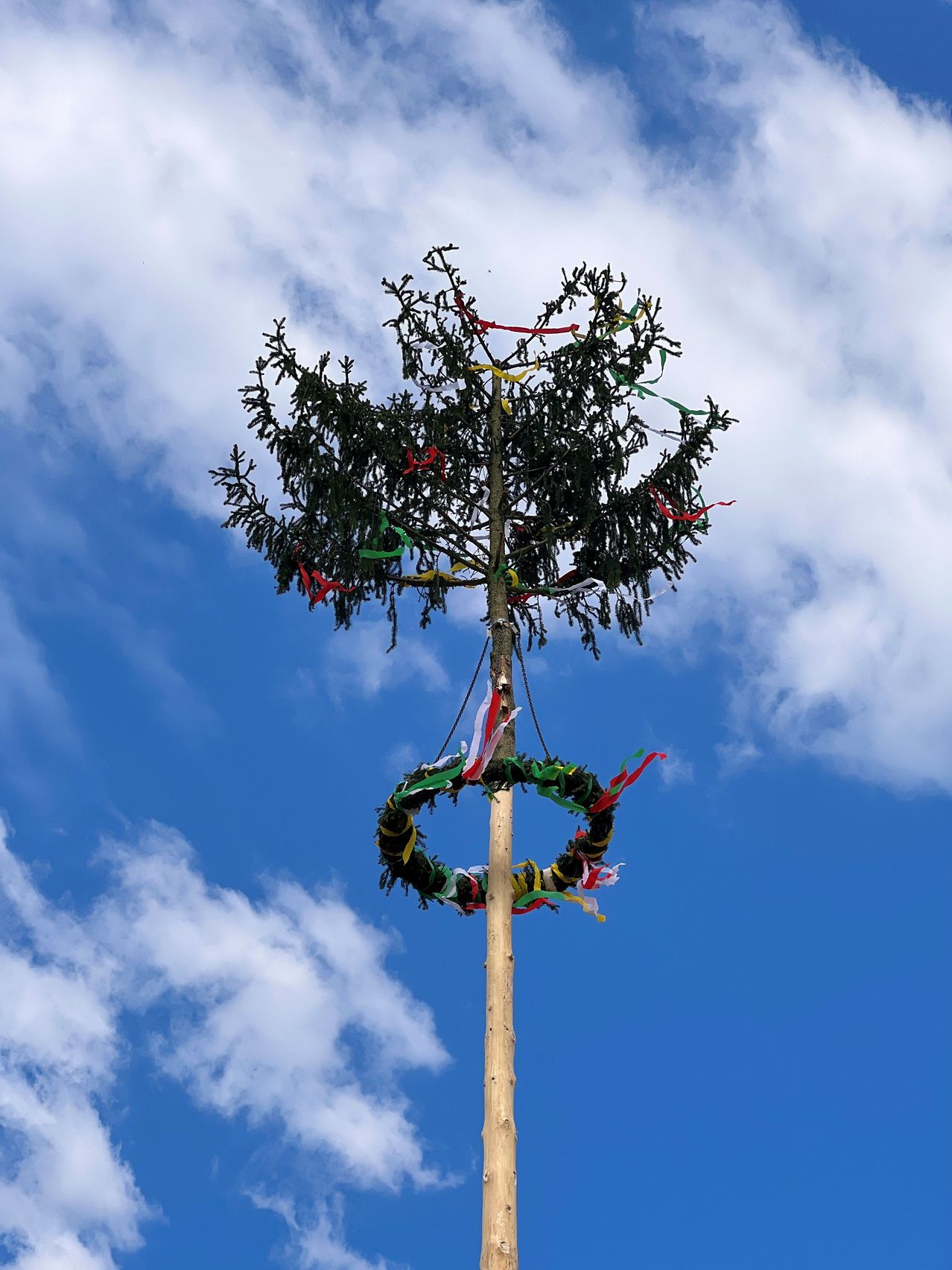 Ein geschmückter Tannenbaum mit bunten Bändern, die daran hängen, steht hoch gegen einen blauen Himmel mit verstreuten Wolken.