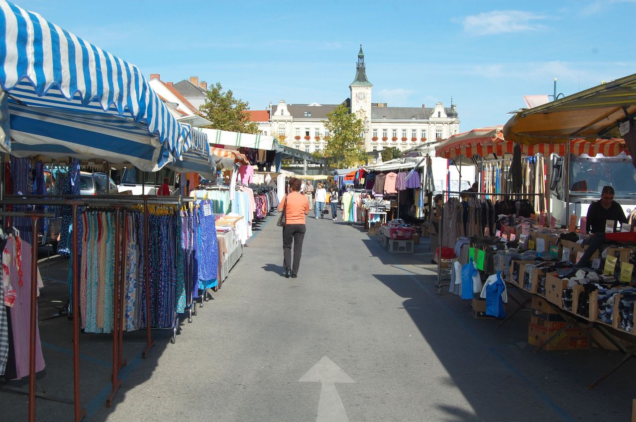 Ein belebter Straßenmarkt mit Kleidungsständen. Eine Frau in Orange geht auf der Straße, während andere stöbern. Ein Gebäude mit Uhrenturm im Hintergrund.