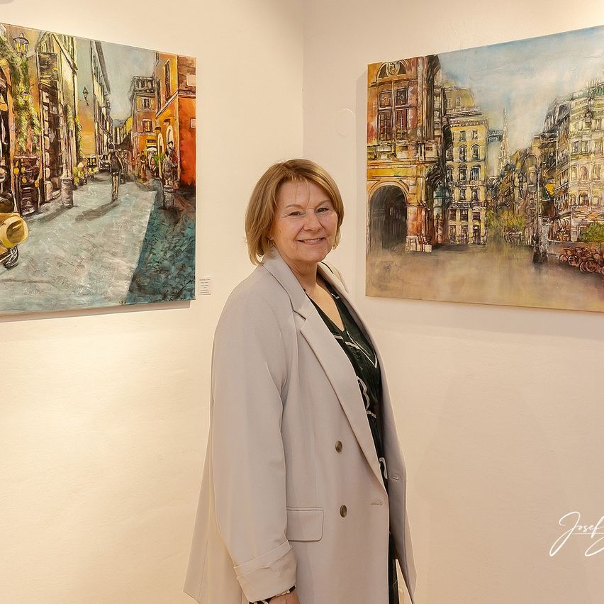 Eine Frau steht in einer Kunstgalerie mit zwei Stadtlandschaftsgemälden, eines zeigt eine europäische Straße und das andere eine Stadtansicht mit Gebäuden und einem Bogen.