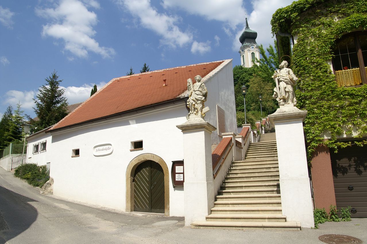 Ein weißes Gebäude mit rotem Dach hat eine Holztür und zwei Statuen. Stufen führen zum Eingang mit zwei Statuen auf beiden Seiten. Im Hintergrund befinden sich ein Turm und Bäume.