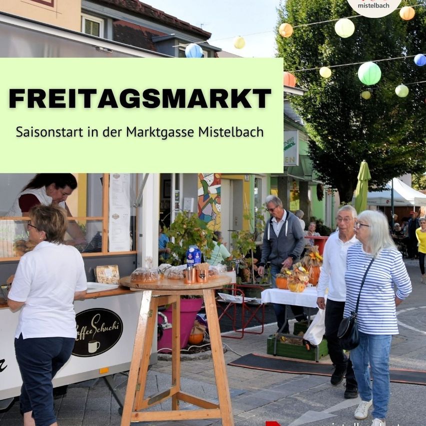 Menschen schlendern auf einer Marktstraße in Mistelbach, Österreich. Stände sind mit Waren aufgestellt, und eine Frau bereitet Essen in einem Food-Truck zu. Bunte Luftballons hängen über ihnen.