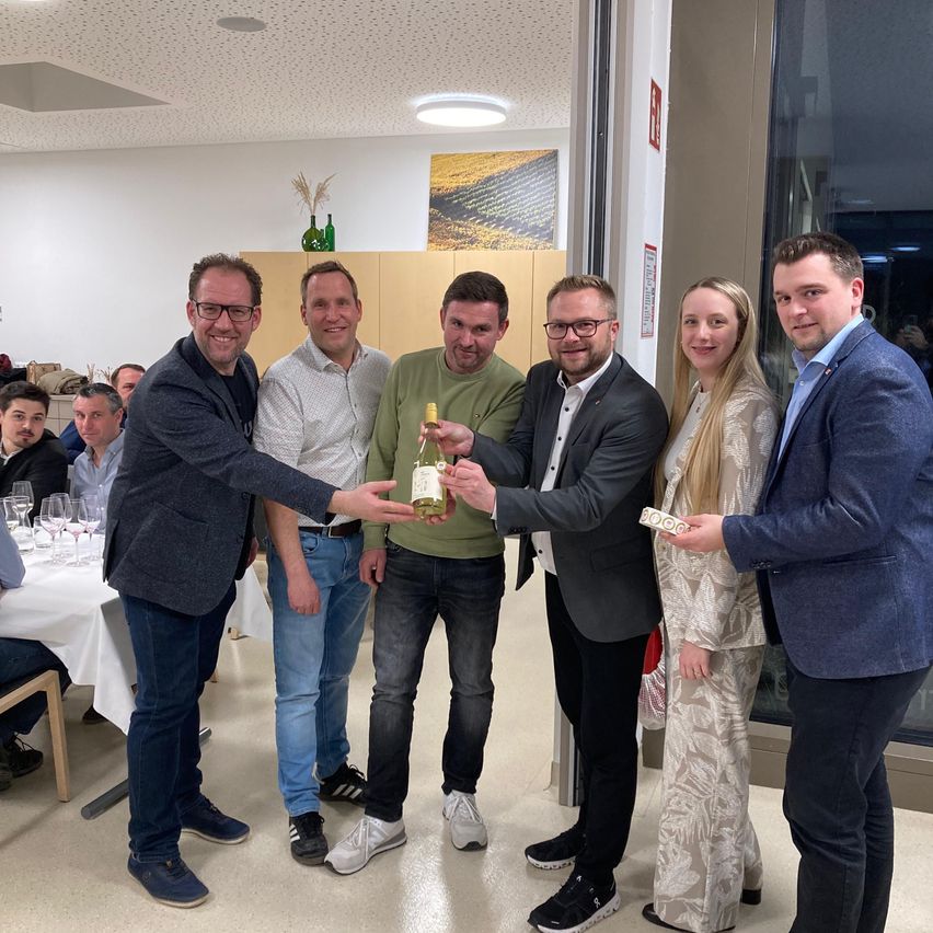 Eine Gruppe Erwachsener steht in einem Raum mit Weingläsern auf einem Tisch und hält eine Flasche Wein und andere Gegenstände. Sie lächeln für ein Foto.