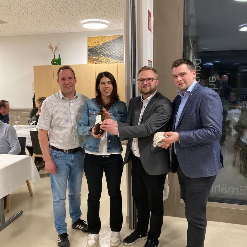 Vier Personen stehen lächelnd in einem Restaurant. Eine Frau hält eine Flasche Wein. Hinter ihnen ist eine Tür geöffnet, und ein Mann macht ein Foto. Im Hintergrund sitzen Menschen an Tischen.