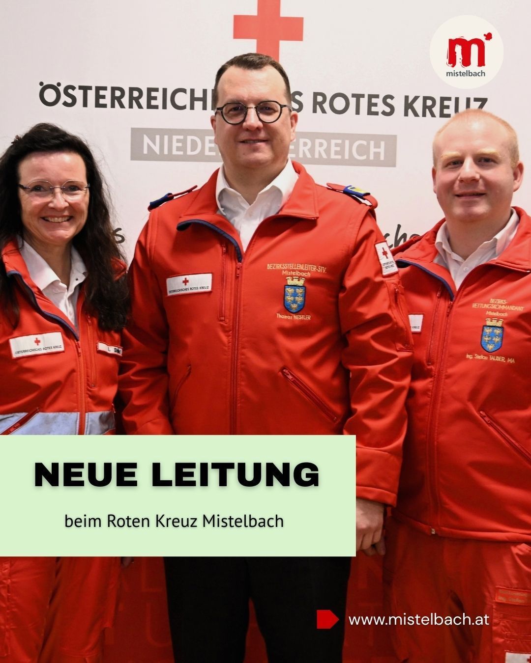 Drei Personen in roten Uniformen posieren für ein Foto vor einem Hintergrund mit 'Rotes Kreuz Mistelbach'. Sie stehen eng beieinander, lächeln und einer trägt eine Brille und einen Abzeichen.
