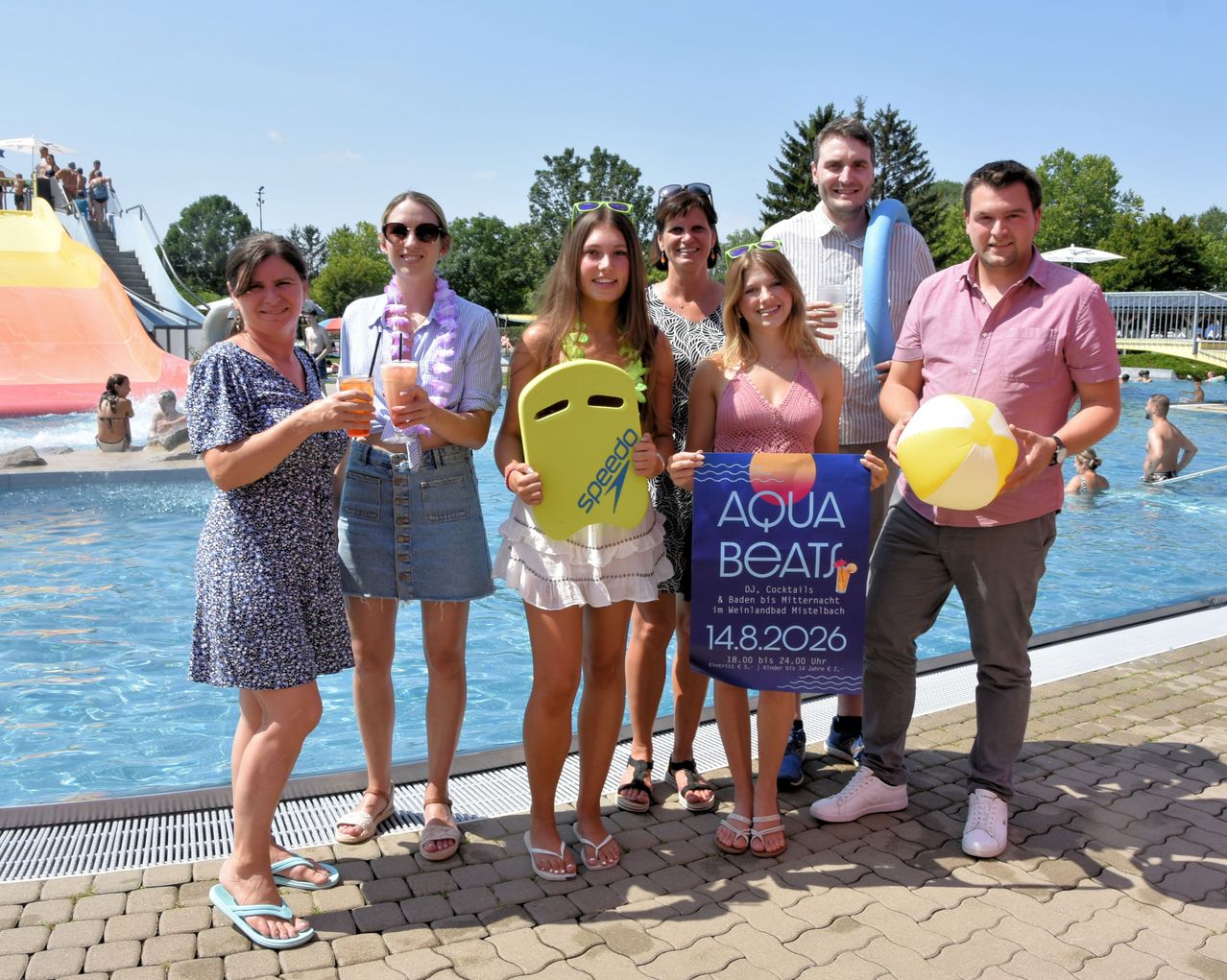 Eine Gruppe von Menschen steht am Pool und lächelt. Sie halten ein Schild für Aqua Beats. Sie tragen lässige Sommerkleidung und einige haben Strandzubehör.