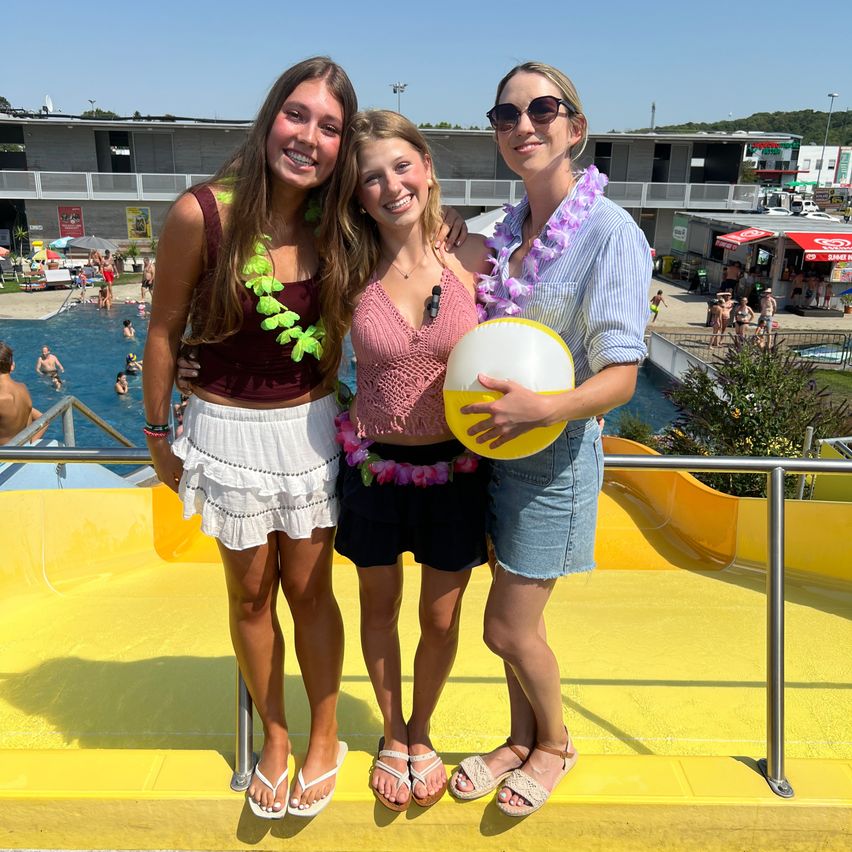 Drei Frauen stehen gemeinsam in einem Wasserpark, eine hält einen Strandball, mit einem Pool und einer Wasserrutsche im Hintergrund.