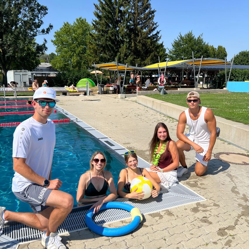 Eine Gruppe von Menschen posiert für ein Foto an einem Schwimmbad. Eine Person hält einen Ball, und andere sitzen oder knien am Rand des Beckens.