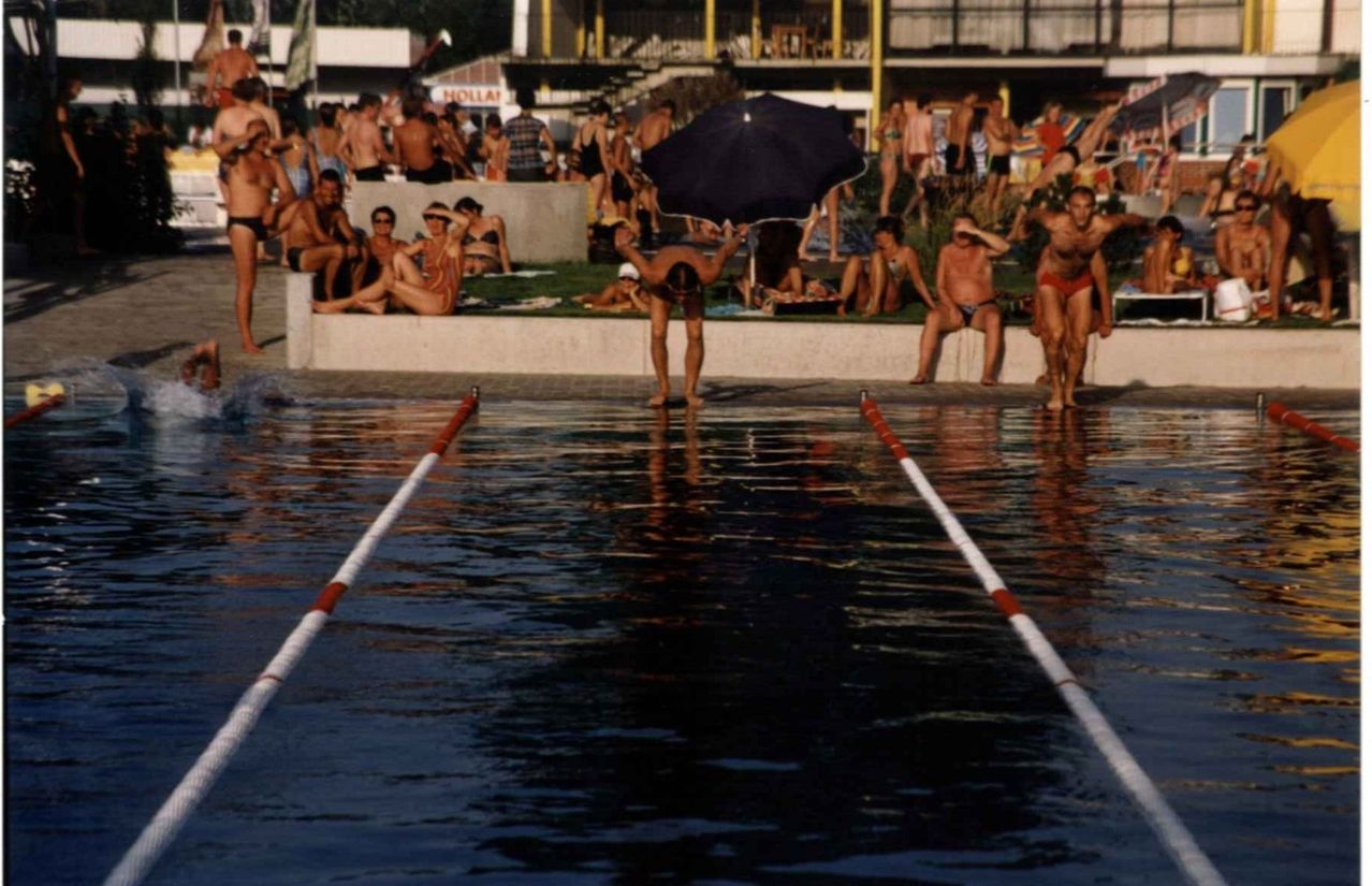 Ein überfüllter Außenpool mit schwimmenden, sitzenden und stehenden Menschen. Ein Mann taucht in den Pool, während andere zusehen.