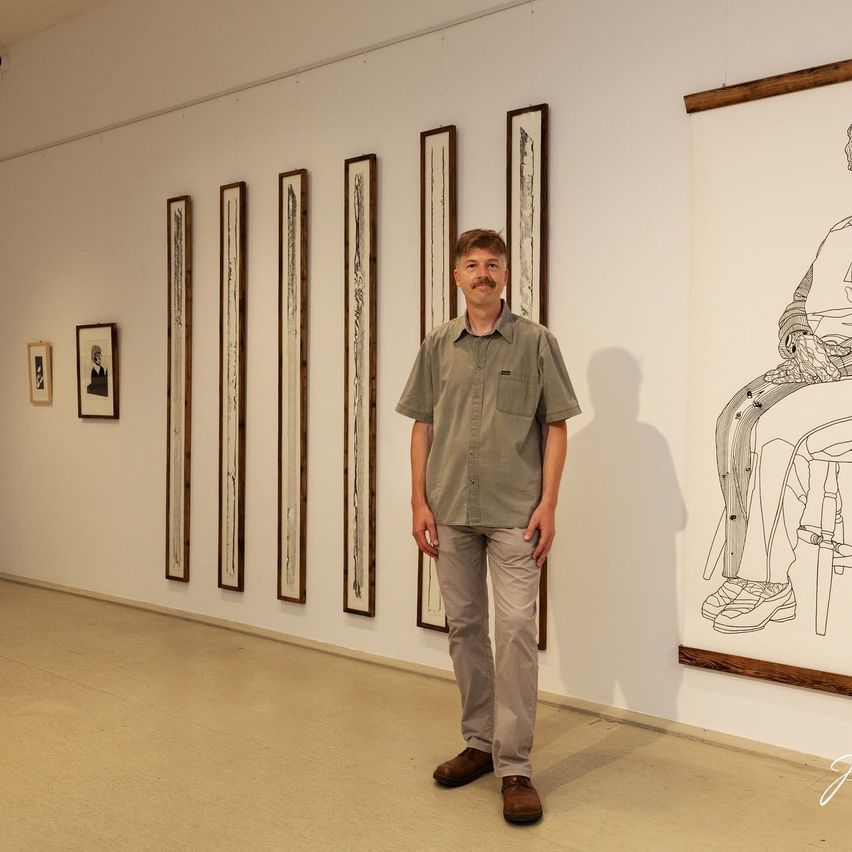 Ein Mann steht in einer Kunstgalerie und sieht eine Wand mit mehreren gerahmten Stücken und einer großen Zeichnung einer sitzenden Person.