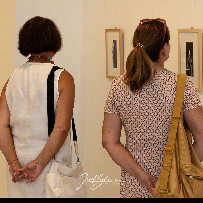 Zwei Frauen stehen vor einer Wand mit gerahmten Bildern, eine trägt ein weißes Kleid und die andere ein gemustertes Kleid und eine braune Tasche.