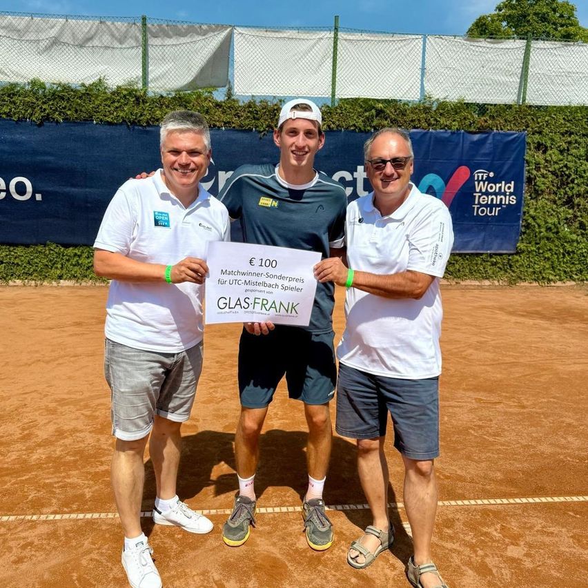 Three men stand on a tennis court, with one holding a certificate for 100 euros. The man in the middle wears a cap and a dark shirt, while the others wear white shirts and shorts. Behind them is a blue banner with the text 'World Tennis Tour'.