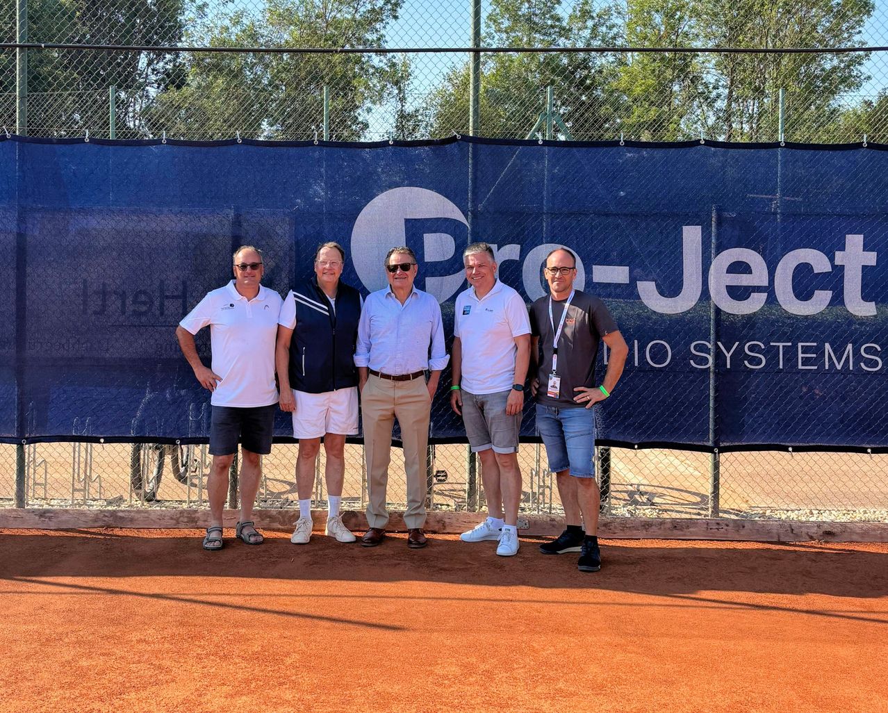 Five men stand in front of a blue banner with the text 'Pro-Ject' and a logo, wearing casual attire and posing for a photo on a tennis court.