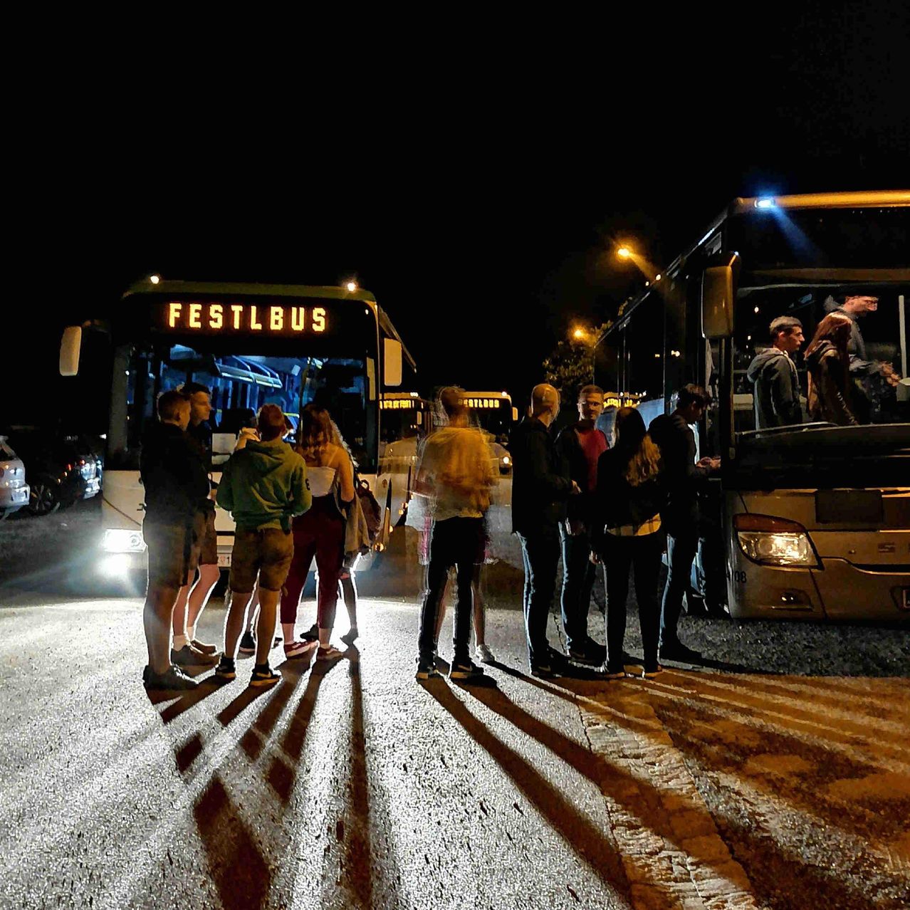Eine Gruppe von Menschen steht nachts in einer Schlange, um in einen Bus namens Festlbus einzusteigen. Der Bus ist am Straßenrand geparkt, und die Scheinwerfer sind eingeschaltet.