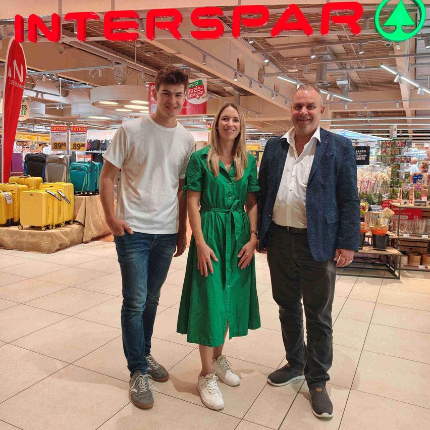 Drei Personen stehen vor einem Interspar-Geschäft und lächeln für ein Foto. Der Mann links trägt ein weißes T-Shirt und Jeans, die Frau in der Mitte trägt ein grünes Kleid und der Mann rechts trägt einen blauen Blazer.