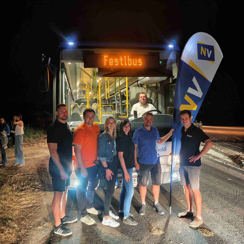 Eine Gruppe von Menschen steht vor einem Bus mit der Aufschrift FestiBus bei Nacht. Der Busfahrer ist drinnen, und eine Fahne mit den Buchstaben NV wird von einer der Personen gehalten.