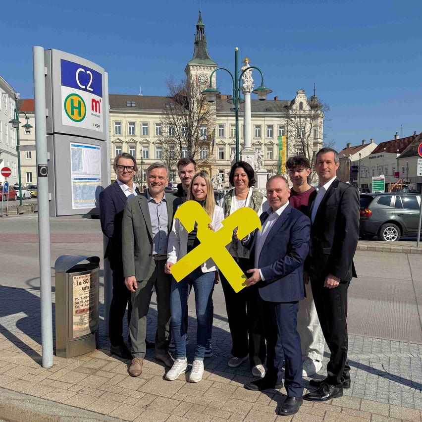 Eine Gruppe von Menschen steht auf einem Bürgersteig, lächelt und posiert für ein Foto mit einem großen gelben X-Zeichen im Vordergrund. Dahinter befindet sich eine Bushaltestelle mit einem Schild und einem Glockenturm in der Ferne.