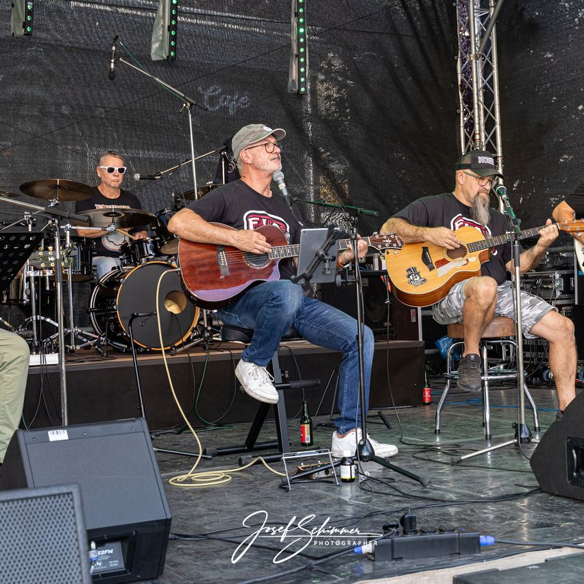 Eine Band spielt auf der Bühne, ein Mann sitzt und spielt Gitarre, ein anderer Mann sitzt und spielt Gitarre, und ein Schlagzeuger im Hintergrund.