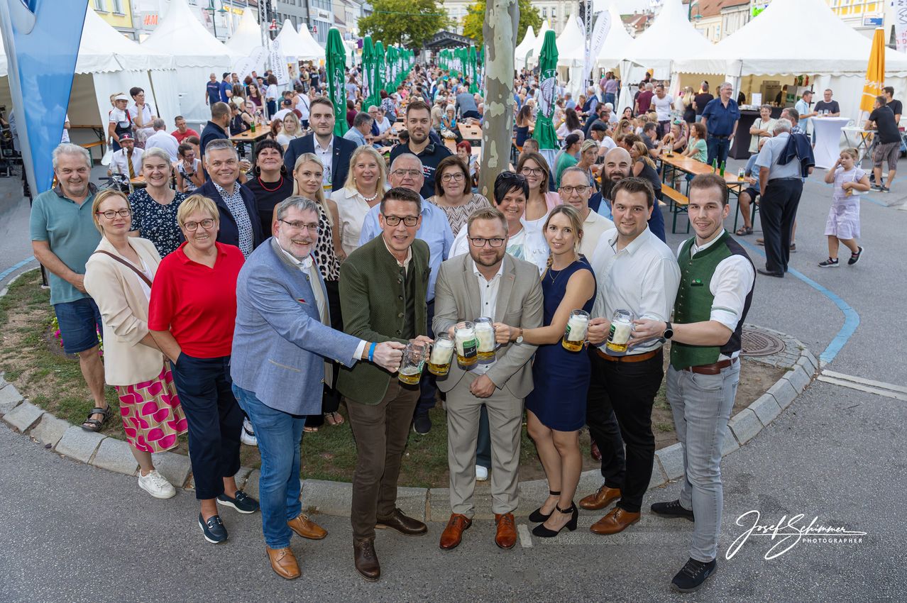 Eine Gruppe von Menschen, darunter Männer und Frauen, steht im Kreis und hält Biergläser in der Hand, wahrscheinlich bei einem Festival oder einer Outdoor-Veranstaltung. Sie lächeln und posieren für ein Foto. Dahinter stehen Zelte, Tische und Stühle, an denen viele Menschen sitzen und stehen. Das Gelände ist von Bäumen und Gras umgeben.