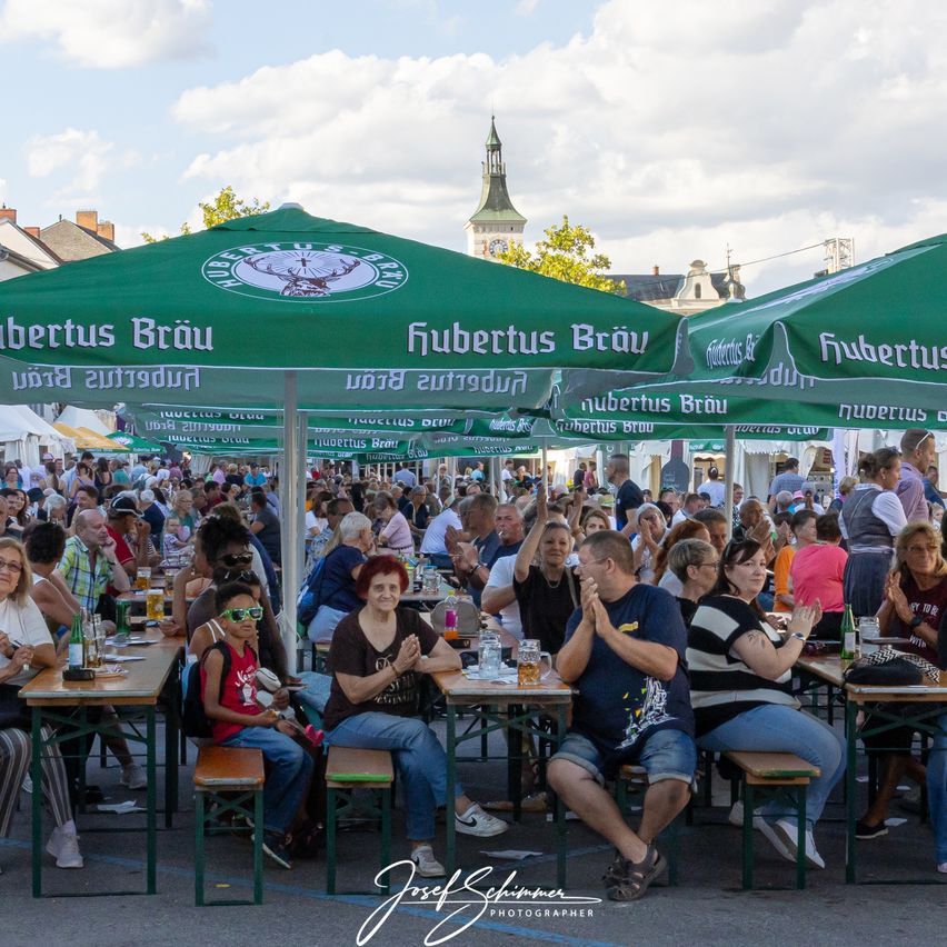 Ein überfüllter Biergarten im Freien mit Menschen unter grünen Schirmen, einige klatschen. Die Tische sind mit Bierkrügen und Essen gefüllt. Im Hintergrund ist ein Turm und ein bewölkter Himmel zu sehen.